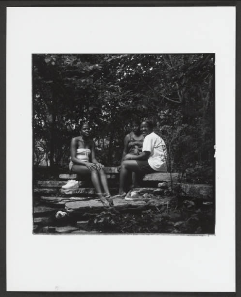 Image: A black and white photograph depicts three young Black teenage girls looking at the camera and casually seated on a low stone wall in a verdant, shadowy wooded area, of Columbus Park. in 1994. Photo Credit: Judith Bromley, Courtesy of Chicago Park District Records: Photographs at the Chicago Public Library. Special Collections & Preservation Division, Digital Link.
